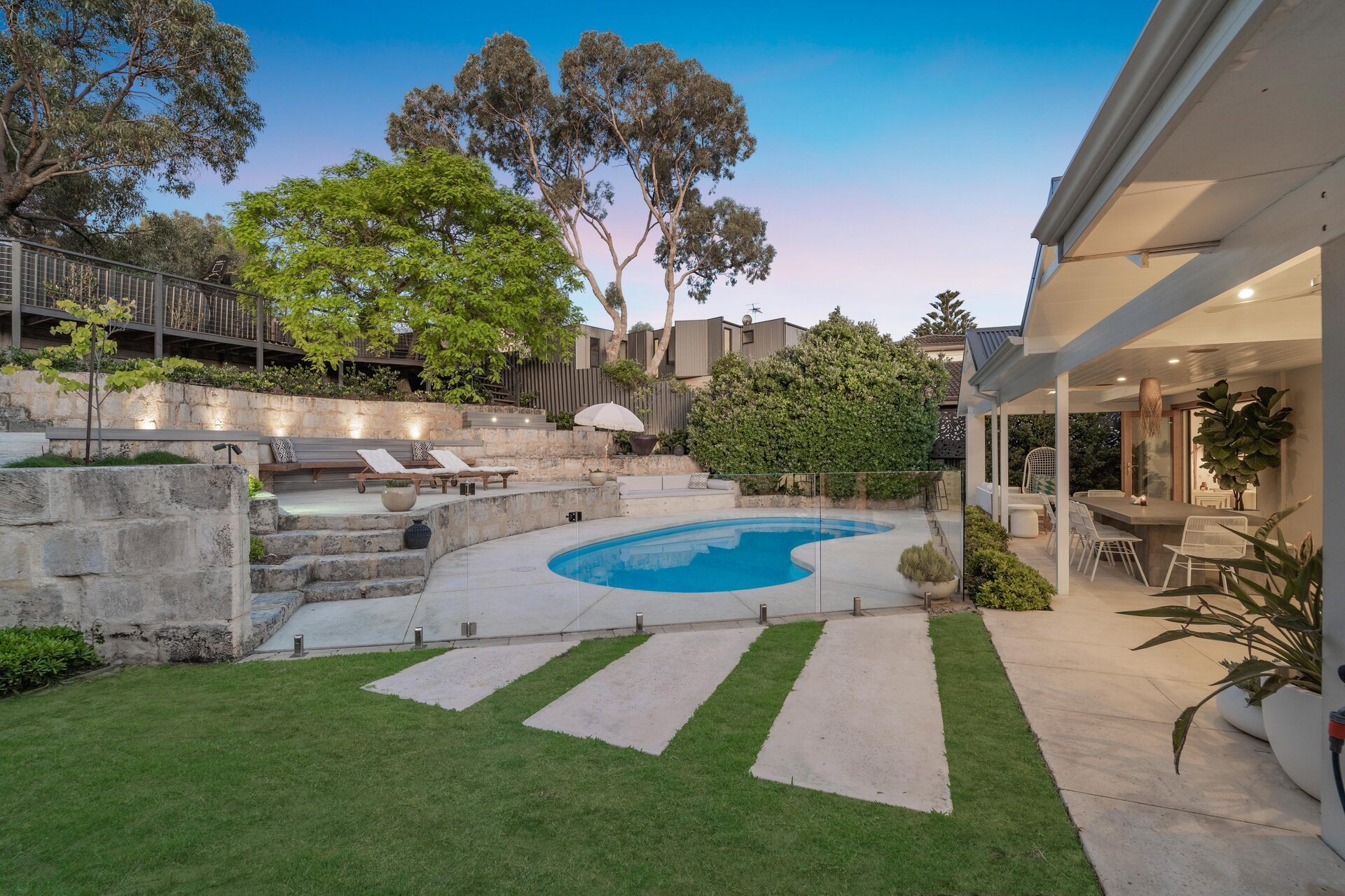 Landscaped backyard at dusk with a swimming pool, limestone terracing and an alfresco area